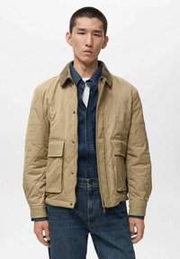 Giacca trapuntata beige con colletto marrone, due tasche frontali e chiusura a bottone, indossata sopra una camicia di denim scuro e jeans blu.