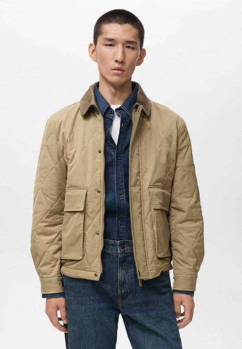 Giacca trapuntata beige con colletto marrone, due tasche frontali e chiusura a bottone, indossata sopra una camicia di denim scuro e jeans blu.