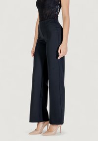 Femme portant un pantalon noir à taille haute et jambes larges, un top en dentelle noire et des escarpins nude à bouts pointus, debout devant un fond uni.