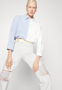 Camisa de botão com a metade esquerda às riscas azuis e a metade direita branca, combinada com jeans rasgados cinza claro e um cinto bege com fivela redonda.