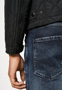 Schwarze gesteppte Jacke mit Reißverschluss und Druckknöpfen, kombiniert mit dunkelblauen Jeans aus Denim, die eine Gesäßtasche und sichtbare Nähte aufweisen.