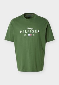 Πράσινο κοντομάνικο t-shirt με στρογγυλή λαιμόκοψη, με την επιγραφή "Tommy Hilfiger 1985" και το κόκκινο, λευκό και μπλε λογότυπο σημαίας του brand μπροστά.