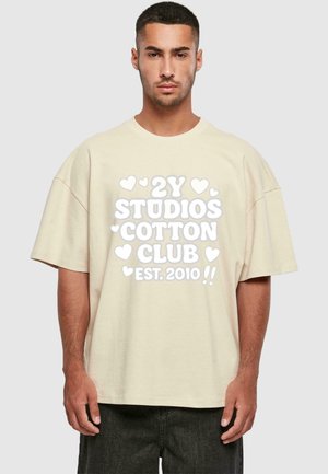 Beige übergroßes T-Shirt aus Baumwolle mit kurzen Ärmeln. Es hat einen großen weißen Aufdruck: "2Y STUDIOS COTTON CLUB EST. 2010" mit dekorativen Herzen.
