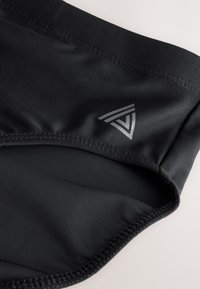 Detail čiernej tkaniny s vystuženým šitím a malým strieborným geometrickým logom na hladkom povrchu.