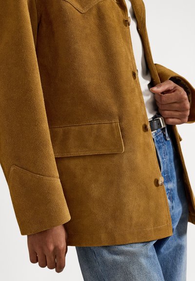 Barna velúrkabátot gombokkal viselő személy, kék farmert és fekete övet viselve, egyik kezével igazítja a kabátot.