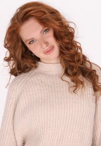 Beige gestrickter Pullover mit hoch geschlossenem Kragen, geripptem Muster und lockerer Passform. Betont lange, lockige Haare, die das Gesicht umrahmen.