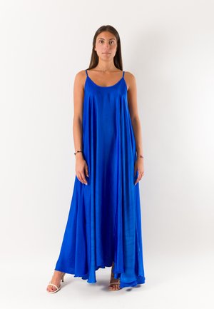 Donna in piedi che indossa un abito lungo senza maniche azzurro brillante e sandali chiari con tacco alto su uno sfondo bianco uniforme.
