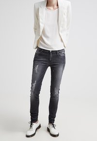 Jean noir slim délavé associé à un blazer blanc texturé et un haut clair ; complété par des chaussures à lacets blanches et noires.