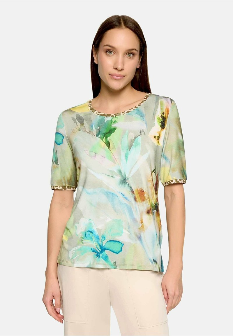 Femme portant un haut à manches courtes imprimé floral aquarelle avec un galon léopard sur l’encolure et les bords des manches, associé à un pantalon beige clair.