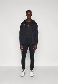 Zwarte sportieve jas met capuchon, zwart aansluitend shirt en zwarte joggingsbroek. Witte sneakers met accenten. Soepele stof, gestroomlijnd ontwerp.