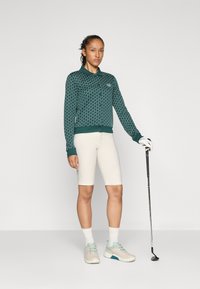 Maglione polo a maniche lunghe verde a fantasia, pantaloni corti crema e scarpe da golf chiare; tiene in mano un bastone da golf; sfondo minimale.