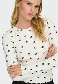 Biała, prążkowana bluzka z długim rękawem z rozrzuconymi czarnymi haftami kwiatowymi i zielonymi liśćmi; falbankowe mankiety dodają tekstury i detali.