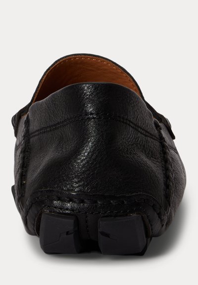 Μαύρο δερμάτινο παπούτσι τύπου μοκασίνι, χωρίς κορδόνια, εμφανιζόμενο από πίσω, που δείχνει την υφή του πάνω μέρους, τις λεπτομέρειες ραφής και τα λαστιχένια πέλματα στη φτέρνα.