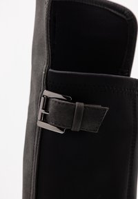 Calzado negro con una parte superior de suave ante y una sección elástica negra en contraste. Incluye un detalle de hebilla metálica y costuras decorativas.