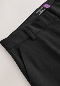 Pantalon noir sur mesure avec passants de ceinture et étiquette de taille Hourglass visible à l'intérieur de la ceinture, sur un fond neutre.