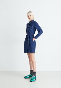 Denim dress with a collar, long sleeves, and a belted waist. Features two chest pockets and a knee-length cut. Worn with green sneakers.