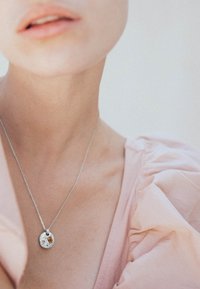 Collana con pendente in argento, caratterizzata da un ciondolo rotondo texture e un piccolo accento in ambra, indossata su una catena contro un tessuto rosa chiaro.