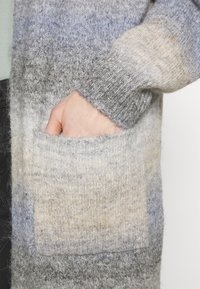 Main partiellement insérée dans la grande poche avant d'un pull ou cardigan en laine tricotée gris et beige.