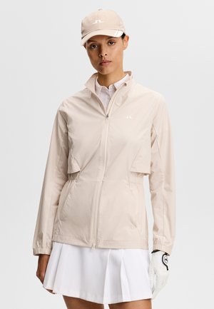 Kvinde iført en beige lynlåstrøje, matchende kasket, hvid polo, plisseret hvid nederdel og hvid golfhandske på højre hånd mod ensfarvet baggrund.