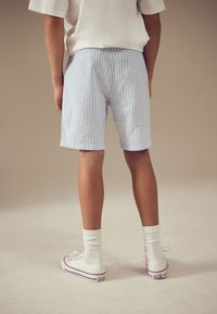 Svetlo modri hlačniki z belimi črtami in sproščenim krojem, kombinirani z belimi visokimi supergami in rebrastimi belimi nogavicami, prikazani od zadaj.