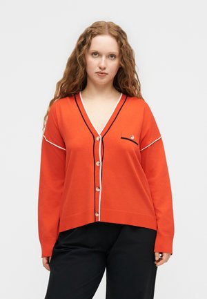 Femeie cu păr lung și creț, purtând un cardigan portocaliu cu nasturi, cu margini negre și albe, și pantaloni negri, stând în picioare pe un fundal simplu.