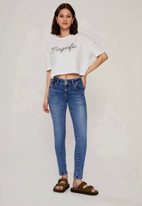 T-shirt branca cropped com "Magnific" impresso em preto, combinada com jeans skinny azuis e sandálias verdes de slide; tecido suave e design ajustado.