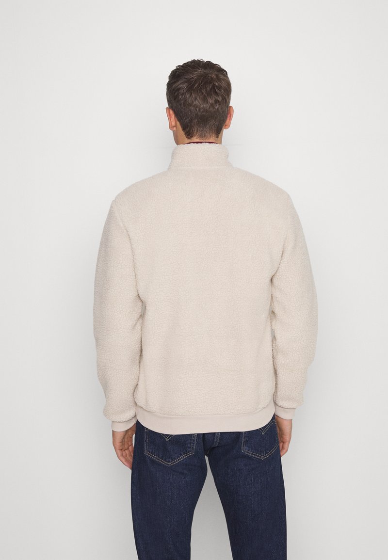 Beiger Fleece-Pullover mit hohem Kragen, strukturiertem Finish und gerippten Bündchen. Kombiniert mit dunkelblauen Jeans.