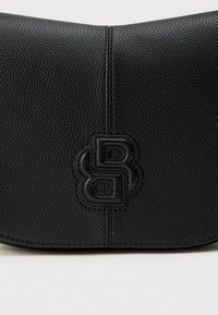 Čierna kožená taška s textúrovaným povrchom, zaobleným tvarom a vyrazeným logom v strede. Má minimálne šitie pozdĺž okrajov.