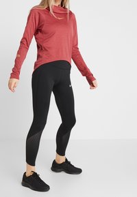 Red long-sleeve top with a cowl neck and gold logo, paired with black leggings featuring mesh accents and logo; black athletic shoes.