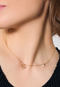 Złoty naszyjnik z łańcuszka z zawieszką w kształcie serca, wygrawerowaną napisem "Mama" oraz z małym akcentem perłowym, zawieszony na szyi.