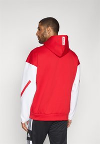 Red and white hoodie with a drawstring hood, featuring a logo on the neck. Smooth fabric, ribbed cuffs, and trim, worn over dark athletic pants.