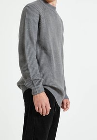 Suéter de punto acanalado gris con un cuello alto, manga larga y aberturas laterales, combinado con pantalones de pana negros.