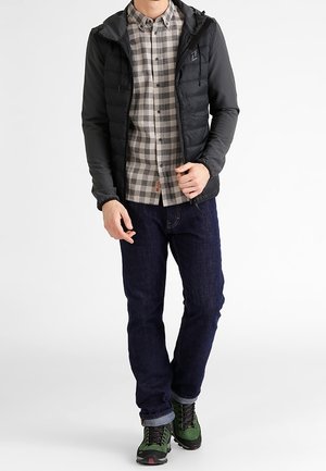 Doudoune noire avec capuche portée sur une chemise à carreaux grise et noire, jean bleu foncé, et chaussures de randonnée vertes avec des accents noirs.