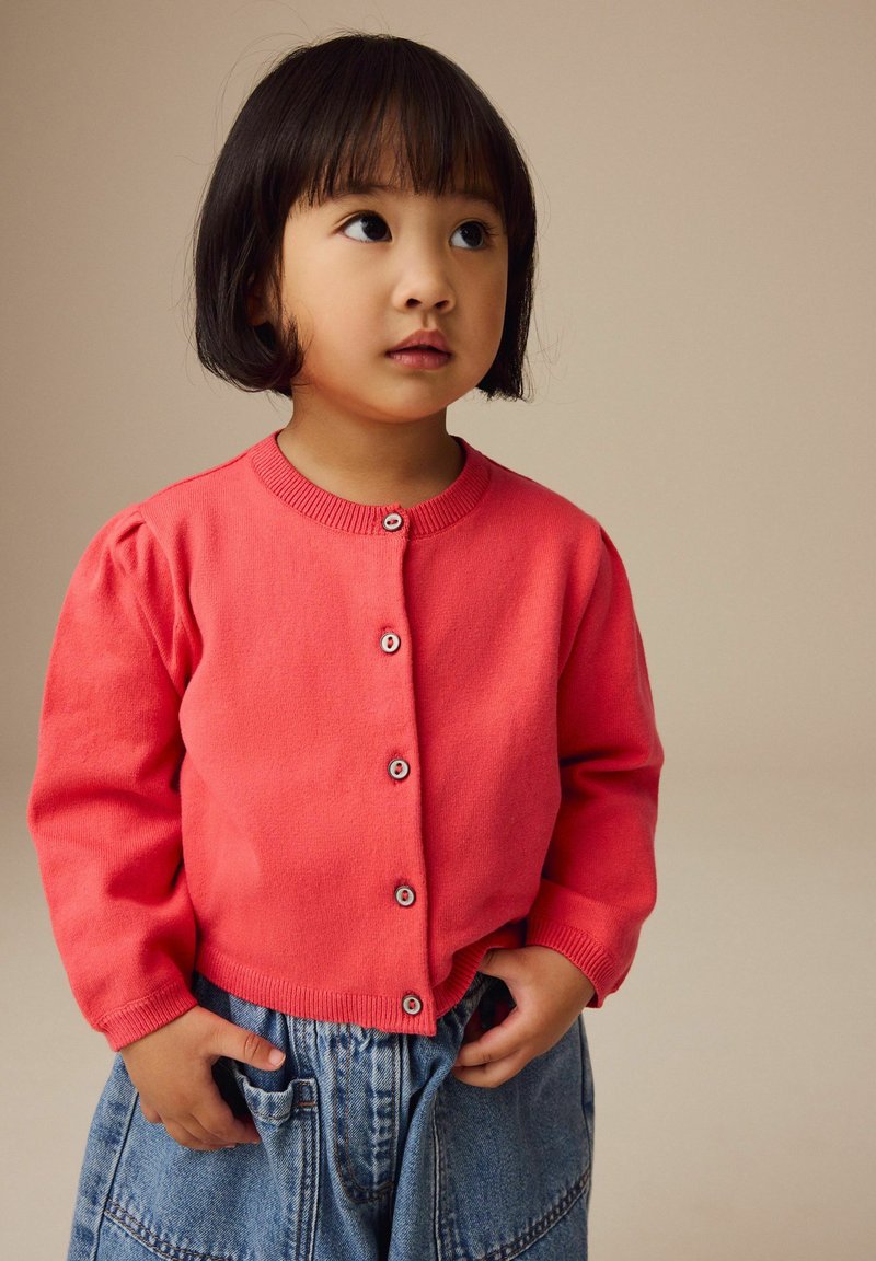 Gilet en corail avec col rond, boutonnière à l'avant et manches à bords côtelés, associé à une jupe en denim clair avec poches et coupe ample.