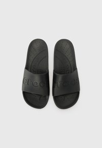 Mustad musta kummist sussid, millel on lai riba koos reljeefse logoga, tekstureeritud tald ventilatsiooniavadega ning lihtne, minimalistlik disain.