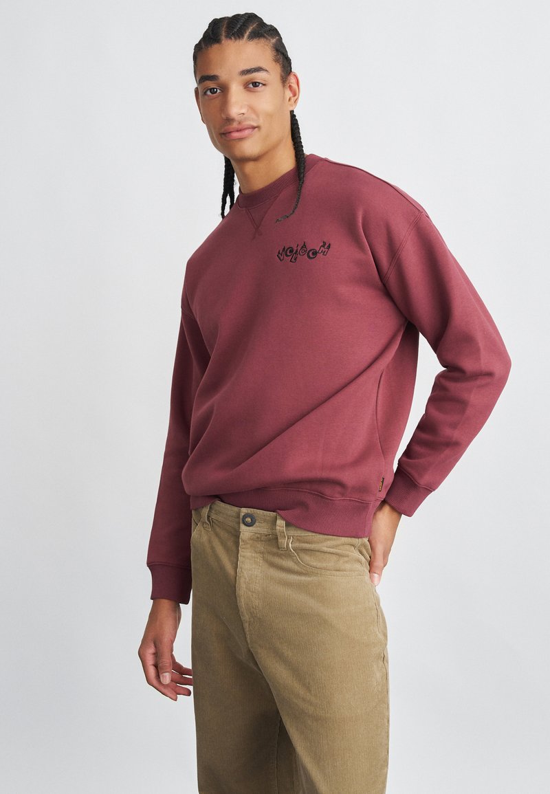 Sweatshirt bordeaux avec logo brodé, col rond et poignets côtelés, associé à un pantalon en velours côtelé beige. Texture lisse, design décontracté.