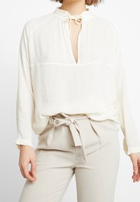 Blusa bianca leggera con colletto arricciato e dettaglio annodato, caratterizzata da una vestibilità morbida e maniche lunghe, abbinata a pantaloni beige a vita alta.