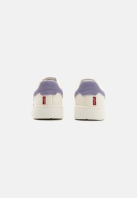 White sneakers with purple suede accents at the heel, featuring textured material and a red logo tab on the side. Thick rubber sole.