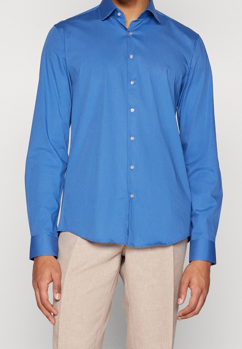 Camicia a maniche lunghe blu realizzata in tessuto morbido, con colletto a punta, chiusura con bottoni e un logo discreto sul petto.