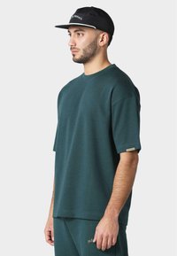 Tmavě zelené oversized bavlněné tričko s krátkými rukávy, kulatým výstřihem a diskrétním logem na rukávu. Nosit s černou kšiltovkou.