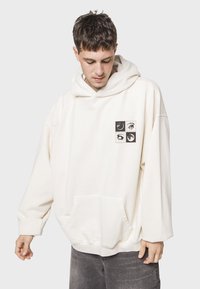 Sudadera de gran tamaño color crema con un bolsillo tipo canguro, que presenta un gráfico impreso con cuatro imágenes cuadradas negras en el pecho.