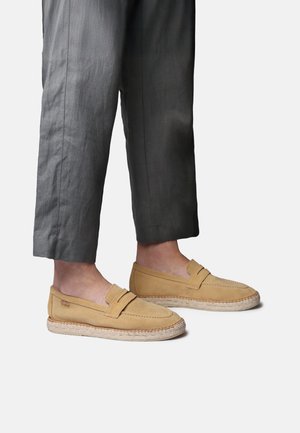 Beżowe zamszowe mokasyny z plecioną podeszwą z juty noszone z szarymi przyciętymi spodniami na jasnym tle.