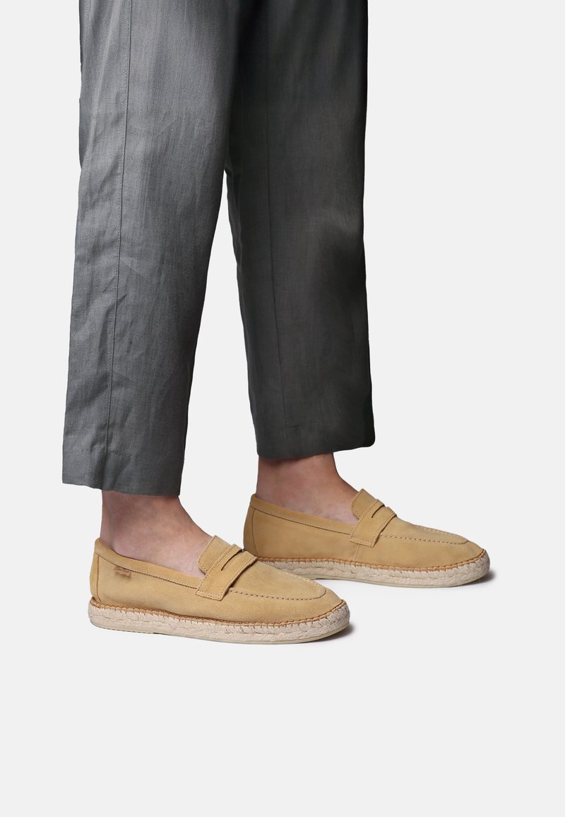 Beežid seemisnahast loafersid punutud juudi tallaga, kantud hallide lühendatud pükstega helel taustal.