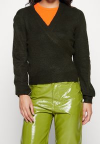 Dunkelgrüner, gestrickter Wickelpullover mit langen Ärmeln, V-Ausschnitt und geripptem Saum, kombiniert mit glänzenden, limonengrünen Hosen.