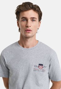 Grijze katoenen t-shirt met korte mouwen en een ronde hals. Kenmerkt zich door een GANT-logo in het marineblauw en rood op de linkerborst.