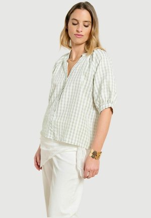 Vrouw draagt een lichtgroene en witte geruite blouse met pofmouwen, witte broek en een gouden armband, kijkt met gesloten ogen naar beneden.