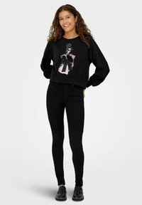Musthave muströöd prindi ja lipsu detailiga must korter rippuv sviiter, millele on lisatud mustad kitsad teksad ja mustad kingad. Kihara juuksed ja kõrvarõngad.