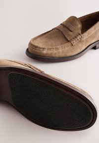 Mocassins en suède marron clair avec un détail cousu, l'un montrant le dessus et l'autre la semelle noire texturée sur un fond uni.