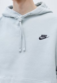 Ljusblå hoodie i mjukt material, med en framficka, dragsnören och en svart broderad Nike-logotyp på bröstet.