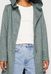 Cappotto verde con texture e cappuccio, indossato sopra una maglietta bianca a coste e jeans chiari a vita alta.